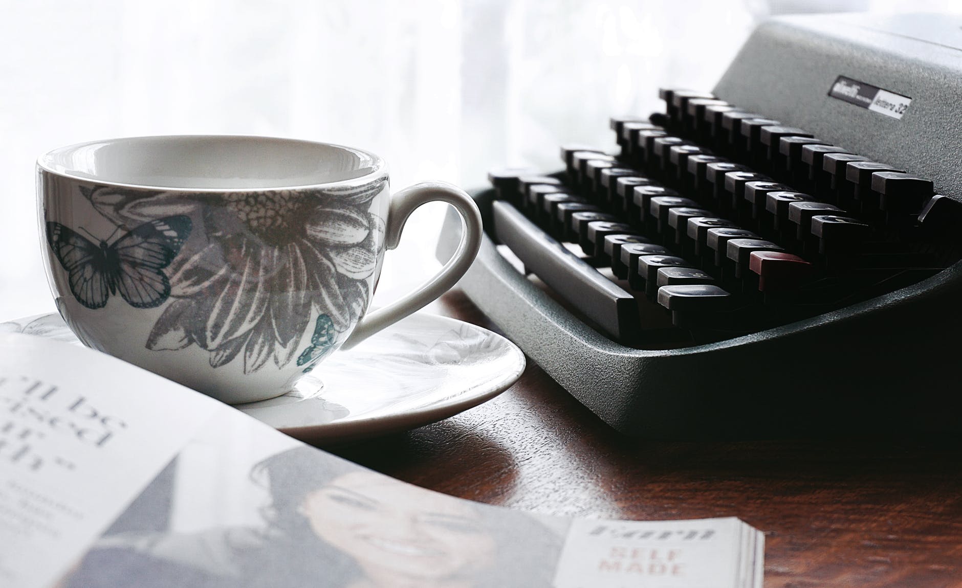 white and gray floral ceramic cup and saucer near black typewriter and book